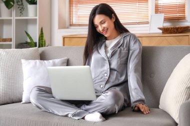Gri pijamalı genç bir kadın evdeki kanepede dizüstü bilgisayarla çalışıyor.