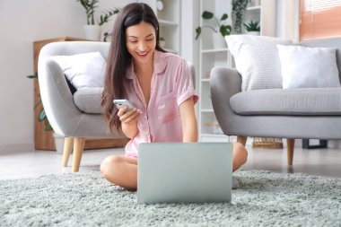 Evde cep telefonu ve dizüstü bilgisayarla çalışan pembe pijamalı serbest çalışan bir kadın.