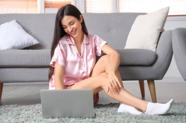 Pembe pijamalı kadın evde dizüstü bilgisayarla halıda çalışıyor.