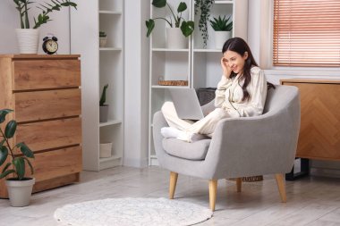 Evdeki koltukta dizüstü bilgisayarla çalışan rahat pijamalı serbest çalışan bir kadın.