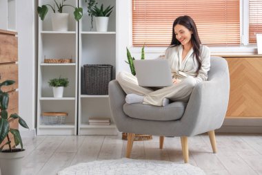 Evdeki koltukta dizüstü bilgisayarla çalışan rahat pijamalı serbest çalışan bir kadın.