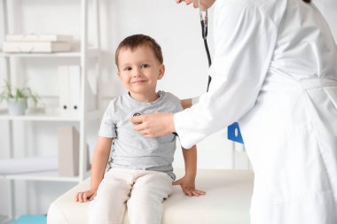 Steteskoplu kadın çocuk doktoru klinikteki kanepede tatlı bir çocuğu dinliyor.