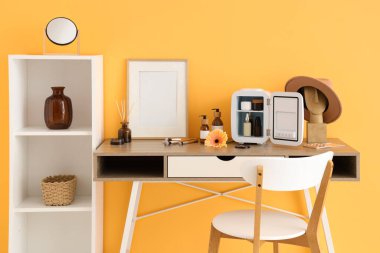 İçinde cilt ürünleri olan küçük kozmetik buzdolabı olan bir masa. Odadaki sarı duvarın yanındaki çerçeve ve çiçek. Yakın çekim.