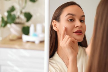 Genç bir kadın banyoda aynanın yanına sivilce bandı yapıştırıyor.