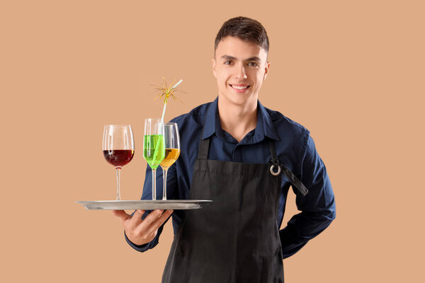 Молодой официант держит поднос с коктейлями на бежевом фоне