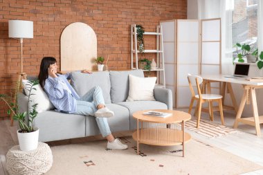 Oturma odasındaki gri koltukta cep telefonuyla konuşan güzel bir kadın.