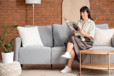 Güzel bir kadın, evdeki gri koltukta dergi okuyor.