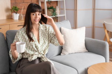 Evdeki koltukta cep telefonuyla konuşan bir fincan çayla güzel bir kadın.