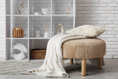 Yastıklı bej pouf, ekose ve raf odası beyaz tuğla duvarın yanında.