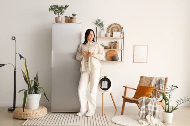 Modern buzdolabının yanında, şık bir mutfakta elinde bir fincan kahveyle duran güzel genç bir kadın.