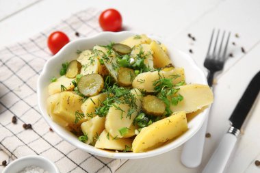 Bir kase taze soğanlı patates salatası ve beyaz ahşap arka planda salatalık turşusu.