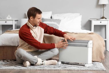 Yatak odasındaki elektrikli konteyner ısıtıcısının yanında ellerini ısıtan genç adam.