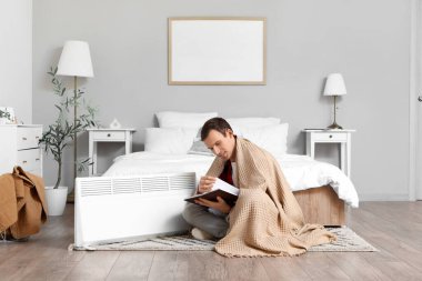 Yatak odasındaki elektrikli ısıtıcının yanında ekoseli kitap okuyan genç adam.