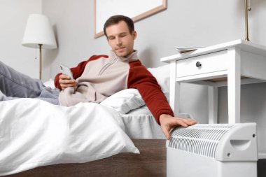 Yatak odasında elektrikli konteyner ısıtıcısının yanında cep telefonu olan genç bir adam yatıyor.