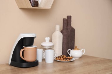 Kahve makinesi, fincanlar, şeker ve kurabiyeler mutfakta bej duvarın yanında.