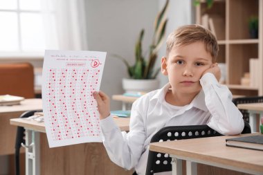 Sınav sonuçlarıyla sınıfta oturan üzgün okul çocuğu.