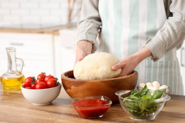 Mutfaktaki ahşap masanın yanında çiğ hamurlu ve pizza malzemeli bir kadın.