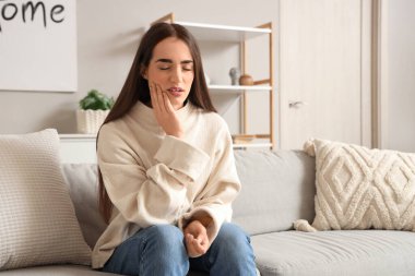 Young woman suffering from toothache on sofa at home clipart