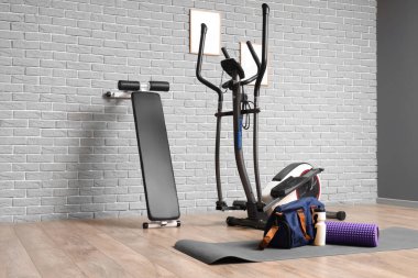 Foam roller, bottle of water, bag on mat and exercisers near grey wall in room clipart