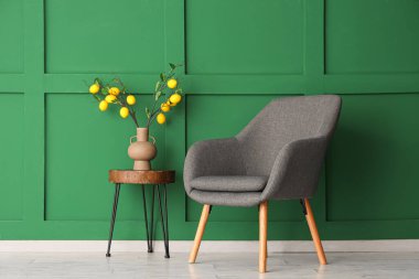 Yeşil duvarın yanındaki sehpanın üzerinde limon ağacı dalları olan gri bir koltuk ve vazo.