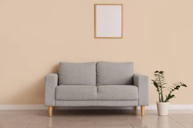 Şık gri kanepe, boş fotoğraf çerçevesi ve bej duvarın yanındaki bitki örtüsü.