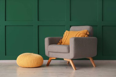 Yeşil duvarın yanında yastıklı ve puflu şık gri koltuk.