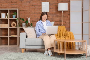 Evdeki kanepede dizüstü bilgisayar kullanan gülümseyen genç kadın.