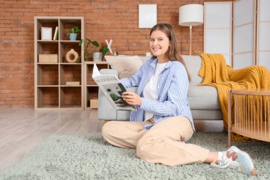 Gülümseyen genç bir kadın evdeki halının üzerinde iç tasarım dergisi okuyor.