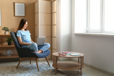 Evdeki koltukta dizüstü bilgisayar kullanan genç bir kadın.