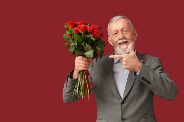 Olgun adam kırmızı arka planda Sevgililer Günü için gül buketini işaret ediyor.