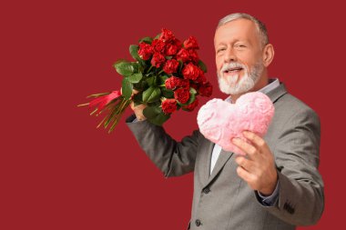 Kırmızı arka planda Sevgililer Günü için gül buketi ve kumaştan kalbi olan olgun bir adam.