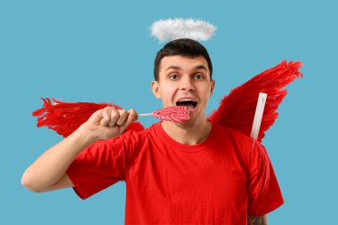 Aşk tanrısı gibi giyinmiş genç bir adam mavi arka planda lolipop yiyor. Sevgililer Günü kutlaması