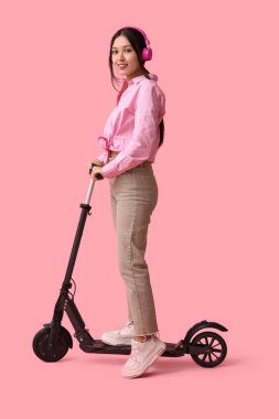 Pembe arka planda modern elektrikli scooterlı kulaklıklı güzel bir Asyalı kadın.