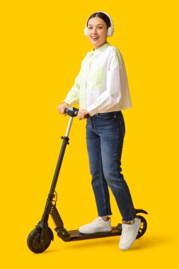 Sarı arka planda modern elektrikli scooterlı kulaklıklı güzel bir Asyalı kadın.