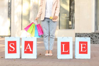 Elinde kredi kartı ve alışveriş torbaları olan genç bir kadın. Şehir caddesindeki giyim mağazasının yakınında.