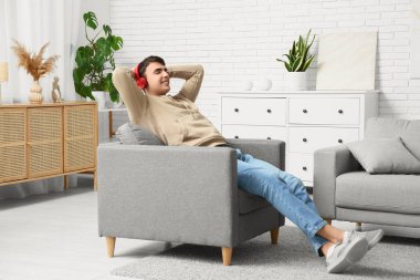 Kulaklıklı yakışıklı genç adam koltukta oturup evde müzik dinliyor.