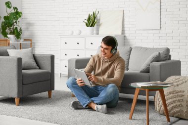 Kulaklığı ve laptopu olan yakışıklı genç adam evde yerde oturuyor.