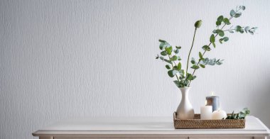 Beyaz duvarın yanındaki masada içinde yeşil okaliptüs bulunan yanan mumlarla dolu bir tepsi. Tasarım için pankart