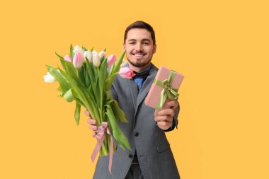 Güzel lalelerden oluşan buketi ve sarı arka planda hediye kutusu olan genç bir adam. Uluslararası Kadınlar Günü