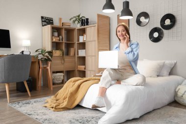 Yatak odasında dizüstü bilgisayarı olan genç bir kadın cep telefonuyla konuşuyor.