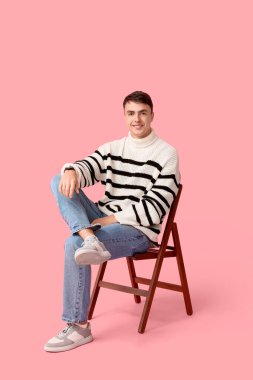Pembe arka planda sandalyede oturan şık çizgili süveterli yakışıklı genç adam.
