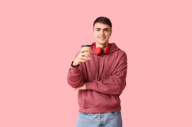 Handsome young man in stylish hoodie with modern headphones and paper cup of hot coffee on pink background clipart