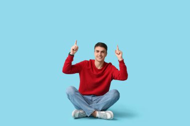 Handsome young man in stylish red sweater pointing at something and sitting on blue background