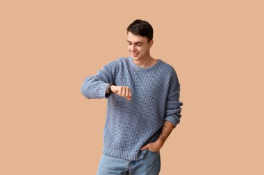 Kahverengi arka planda modern akıllı kol saati olan şık mavi kazaklı yakışıklı bir genç.