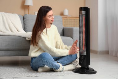 Evde elektrikli ısıtıcının yanında elleri ısıtan genç bir kadın.