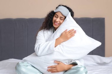 Yatak odasında yumuşak yastıklı pijama ve maske takan güzel Afro-Amerikan kadın. Dünya Uyku Günü