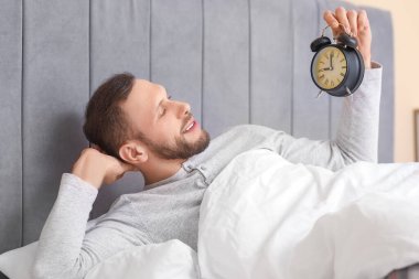 Yatak odasında pijamalı, çalar saati olan genç bir adam yatıyor. Dünya Uyku Günü