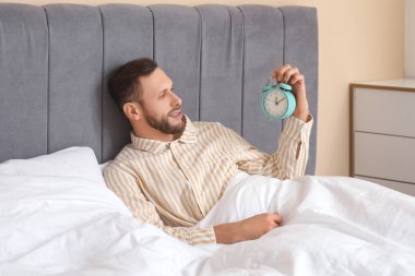 Yatak odasında pijamalı, çalar saati olan genç bir adam yatıyor. Dünya Uyku Günü