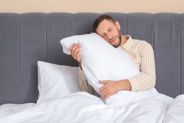 Yatak odasında yumuşak yastıklı pijamalı genç bir adam oturuyor. Dünya Uyku Günü