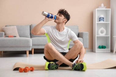 Evde yoga minderinde oturmuş su içen genç bir adam.